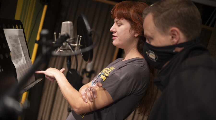 Zdjęcie przedstawia dwóch młodych ludzi konsultujących tekst w studiu nagraniowym. Kobieta ma długie, rude włosy i ubrana jest w koszulkę z nadrukiem. Mężczyzna, ubrany w maskę, podąża za palcem koleżanki, która wskazuje fragmenty tekstu.
