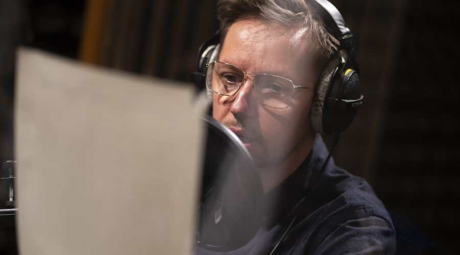 Zdjęcie przedstawia głowę oraz ramiona mężczyzny, w średnim wieku, w okularach, zapatrzonego w kartkę papieru. To Bartek Lis – jedna z osób prowadzących warsztaty. Słuchawki na jego uszach oraz mikrofon w sali wskazują na realizowanie nagrania dźwiękowego.