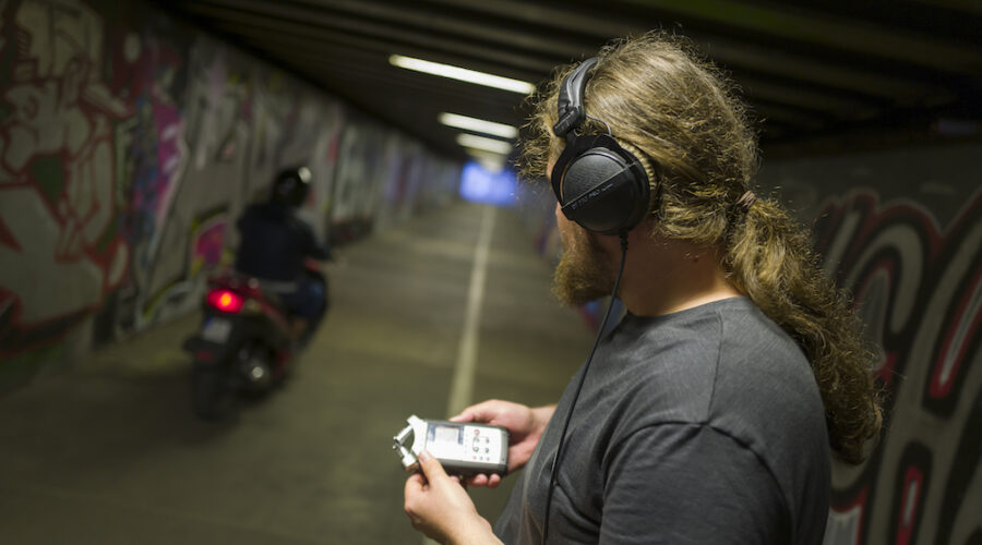 Zdjęcie przedstawia popiersie młodego mężczyzny odwróconego do nas plecami. Chłopak stoi w tunelu. Na uszach ma założone słuchawki, a w dłoniach trzyma dyktafon. Jego głowa skierowana jest w stronę przejeżdżającego właśnie motocykla. Ściany tunelu pomalowane są w różnobarwne graffiti.